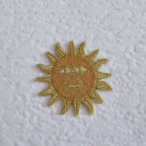 May include: A yellow embroidered sun with a brown face and white eyes. The sun is a patch or iron-on.