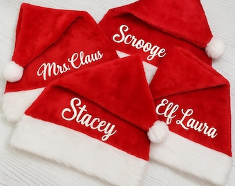 May include: Four red Santa hats with white trim and a white pom-pom. Each hat has a different name in white script: "Mrs. Claus", "Scrooge", "Stacey", and "Elf Laura". The hats are arranged on a light-colored surface.