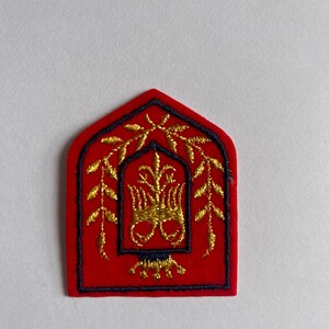 May include: A red felt patch with a gold embroidered design. The design features a stylized plant with leaves and a central motif. The patch is outlined in black thread.