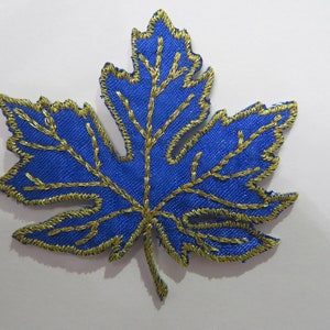 Könnte beinhalten: Ein blaues Stoffahornblatt mit goldener Stickerei. Das Blatt ist mit goldfarbenem Garn umrandet und hat goldene Adern.