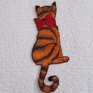 May include: An embroidered iron-on patch of a brown and orange tabby cat with a red bow sitting with its tail curled around its back.