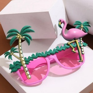 May include: Pink plastic sunglasses with a flamingo and palm trees on the frame. The flamingo is pink with a gold beak and legs. The palm trees are green with gold trunks.