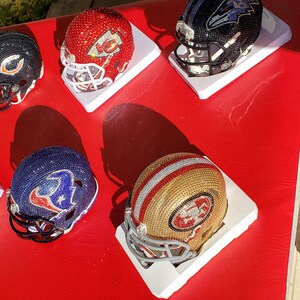 Könnte beinhalten: Sechs Miniatur-Footballhelme, verziert mit Strasssteinen und Teamlogos. Die Helme werden auf weißen Sockeln präsentiert und sind in einer Reihe auf einer roten Oberfläche angeordnet.