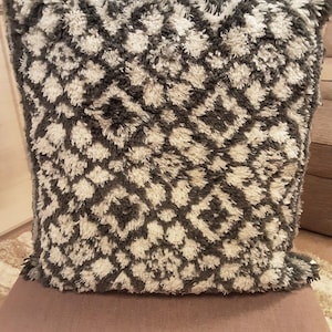 May include: A square cushion with a textured, shaggy design. The cushion features a repeating geometric pattern in shades of grey and white. The cushion is placed on a neutral-coloured surface.