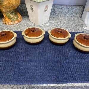 May include: Four round ceramic pots with lids, each featuring a two-tone design with a glossy dark brown top and a smooth cream-colored base. Each pot has two small integrated side handles and a central knob on the lid for easy lifting.