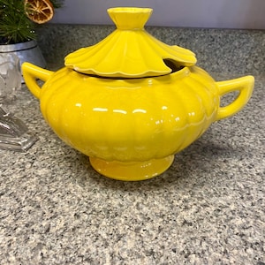 May include: A bright yellow ceramic soup tureen with a lid. The tureen has two handles and a pedestal base. The lid has a decorative, scalloped edge and a small handle. The tureen is likely vintage and adds a pop of color to any kitchen.