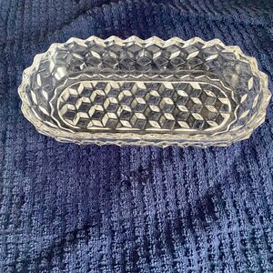 May include: Clear, oval-shaped glass dish with a diamond pattern in the center and a textured, zig-zag edge. The dish is empty and sits on a blue textured surface. This decorative dish is suitable for serving or display.