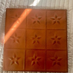 May include: Square, copper-colored decorative tile with a repeating pattern of embossed stars. The tile is divided into nine squares, each featuring a five-pointed star. The surface has a textured, speckled appearance.