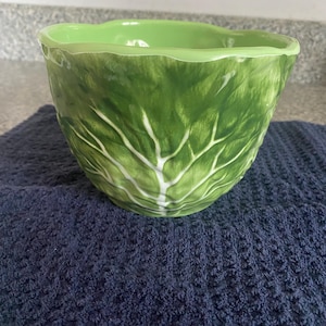 May include: A green ceramic bowl with a cabbage leaf design. The bowl has a wavy rim and a textured exterior, with white veins painted on the green surface. The bowl is sitting on a dark blue towel.