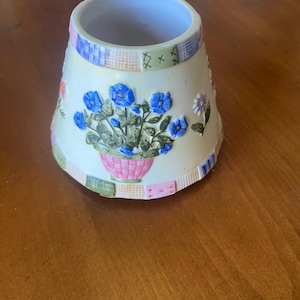 May include: A ceramic candle shade with a tapered, bell-like shape. The exterior features a cream-colored base decorated with embossed blue flowers in a pink pot, surrounded by a patchwork-style border of small, colorful squares and patterns.