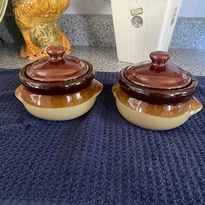 May include: Two small, round ceramic pots with matching lids, featuring a two-tone design with a dark brown glazed upper section and a lighter tan base. Each pot has two small side handles and a central knob on the lid for easy lifting.