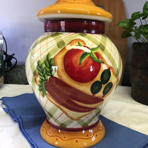 May include: A ceramic jar with a lid, decorated with a painted design of a tomato, olives, and other vegetables on a checkered background. The jar is yellow, green, red, and brown.