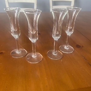 May include: Four clear glass champagne flutes with a tulip-shaped bowl and a slender stem. The flutes are arranged on a wooden table, with a blurred background of white chairs and a light blue wall.
