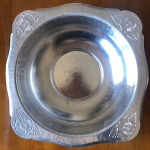 May include: A silver metal bowl with a square shape and a scalloped edge. The bowl has a floral design embossed on the rim.