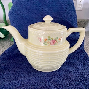 May include: Cream-colored ceramic teapot with a lid and handle. The teapot features a floral design with pink roses and orange flowers. The body of the teapot has a textured, horizontal ribbed pattern. The teapot is sitting on a blue surface.