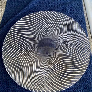 May include: A round, decorative plate with a spiral design. The plate features alternating black and white lines radiating from the center. The plate is resting on a dark blue textured surface.