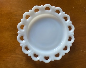 May include: A white, round, vintage plate with a scalloped edge featuring decorative cut-out designs. The plate is made of opaque glass and sits on a wooden surface.