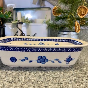 May include: Rectangular ceramic baking dish with a white base and blue floral pattern. The rim features a dotted blue border. The dish has a slightly raised edge and is designed for oven use.