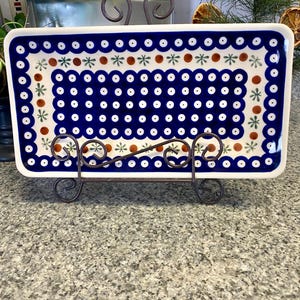 May include: A rectangular ceramic serving dish with a blue and white floral pattern, displayed on a decorative metal stand. The dish features a border of blue dots and a central design of white dots, brown circles, and green star-like shapes.