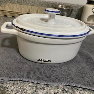 May include: White ceramic Dutch oven with a lid, featuring a blue rim and handle accents. The pot has a distressed, rustic look with brown detailing around the edges. The pot is sitting on a gray towel.
