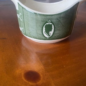 May include: Cream-colored gravy boat with a green patterned design. The boat features a green silhouette of a person in an oval frame. The boat has a curved spout and a wide, open top. The boat is made of ceramic.