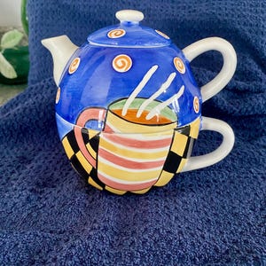 May include: A whimsical ceramic teapot with a matching teacup design. The teapot is primarily blue with orange swirl accents and white lines. The teacup portion features a colorful striped pattern and a black and yellow checkered design.
