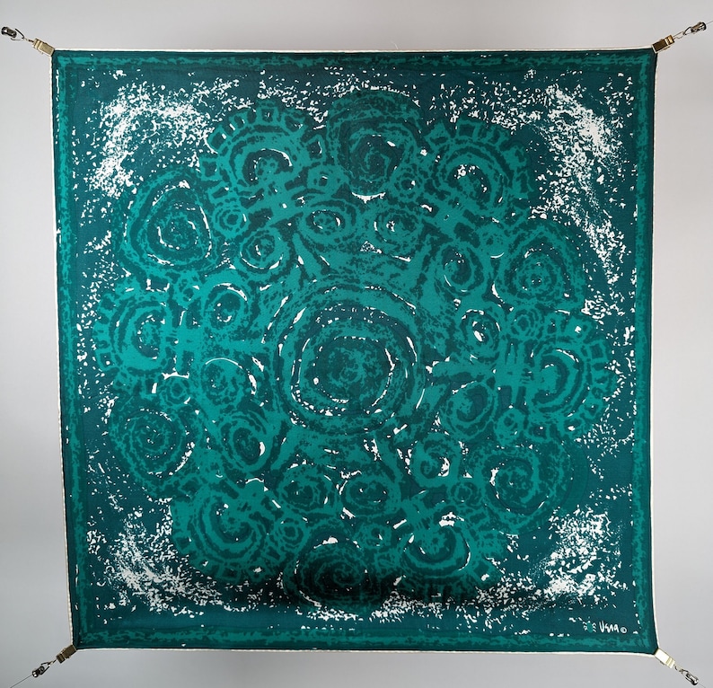 May include: A teal blue square scarf with a white and teal abstract pattern of swirling circles. The scarf has a white border and the signature "V.M.A." in the bottom right corner.