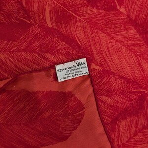 May include: A red silk scarf with a feather pattern. The scarf is hand-rolled and water-repellent. The label reads "scarves by Vina all silk handrolled made in Japan WATER-REPELLENT"