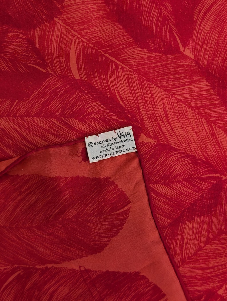 May include: A red silk scarf with a feather pattern. The scarf is hand-rolled and water-repellent. The label reads "scarves by Vina all silk handrolled made in Japan WATER-REPELLENT"