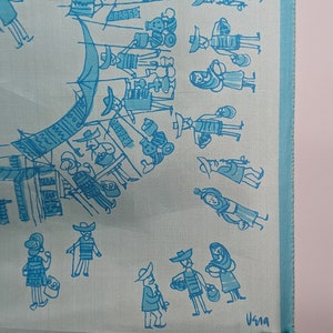 May include: A light blue silk scarf with a blue and white illustration of a market scene. The scarf has a blue border and the text "Usma" is printed in the bottom right corner.