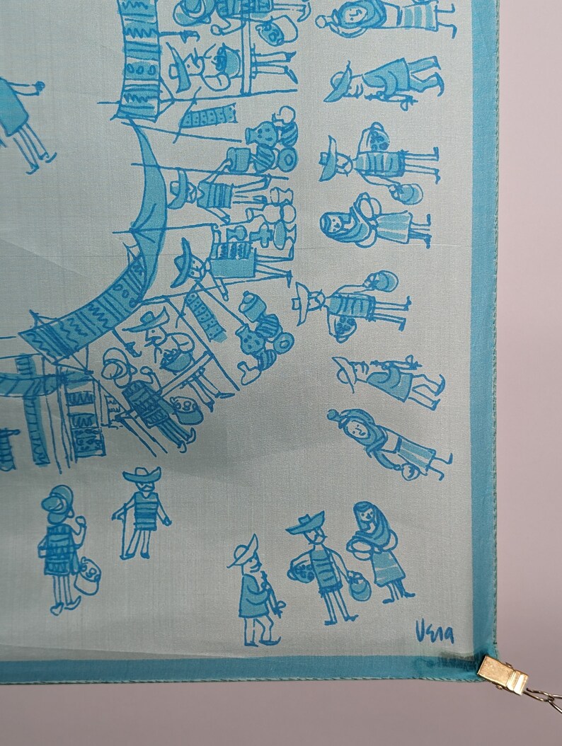 May include: A light blue silk scarf with a blue and white illustration of a market scene. The scarf has a blue border and the text "Usma" is printed in the bottom right corner.