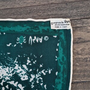 May include: A green silk scarf with a white abstract design and a white border. The scarf has a label that reads "scarves by V&A all silk handrolled made in Japan".