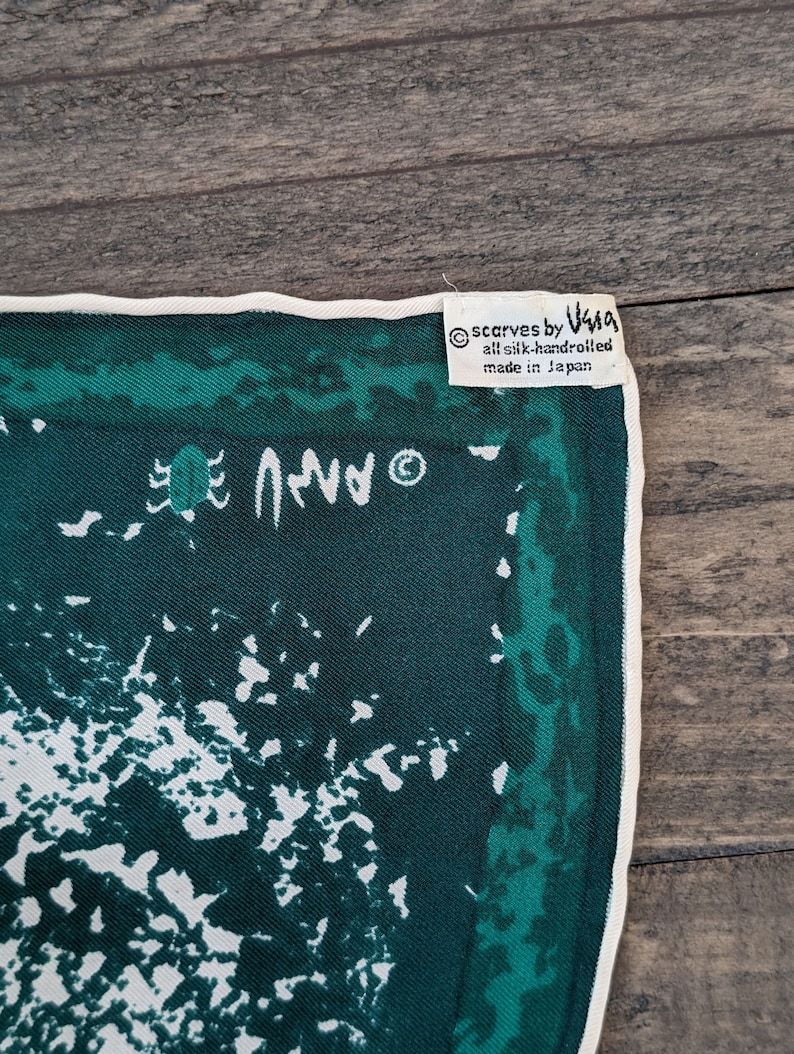May include: A green silk scarf with a white abstract design and a white border. The scarf has a label that reads "scarves by V&A all silk handrolled made in Japan".