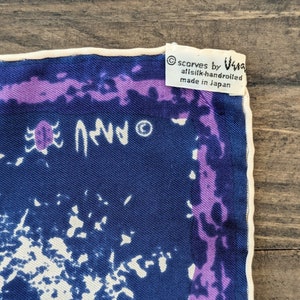 May include: A blue silk scarf with a white and purple abstract design. The scarf has a white border and a label that reads "scarves by Ugg, all silk-handrolled, made in Japan."