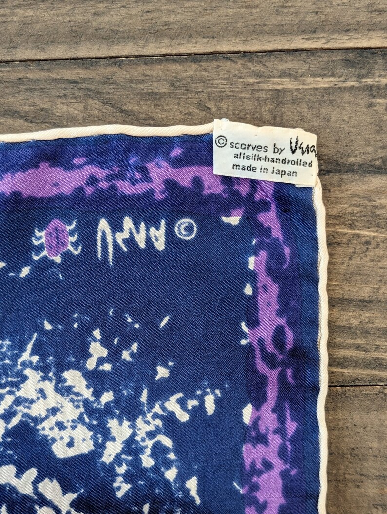 May include: A blue silk scarf with a white and purple abstract design. The scarf has a white border and a label that reads "scarves by Ugg, all silk-handrolled, made in Japan."