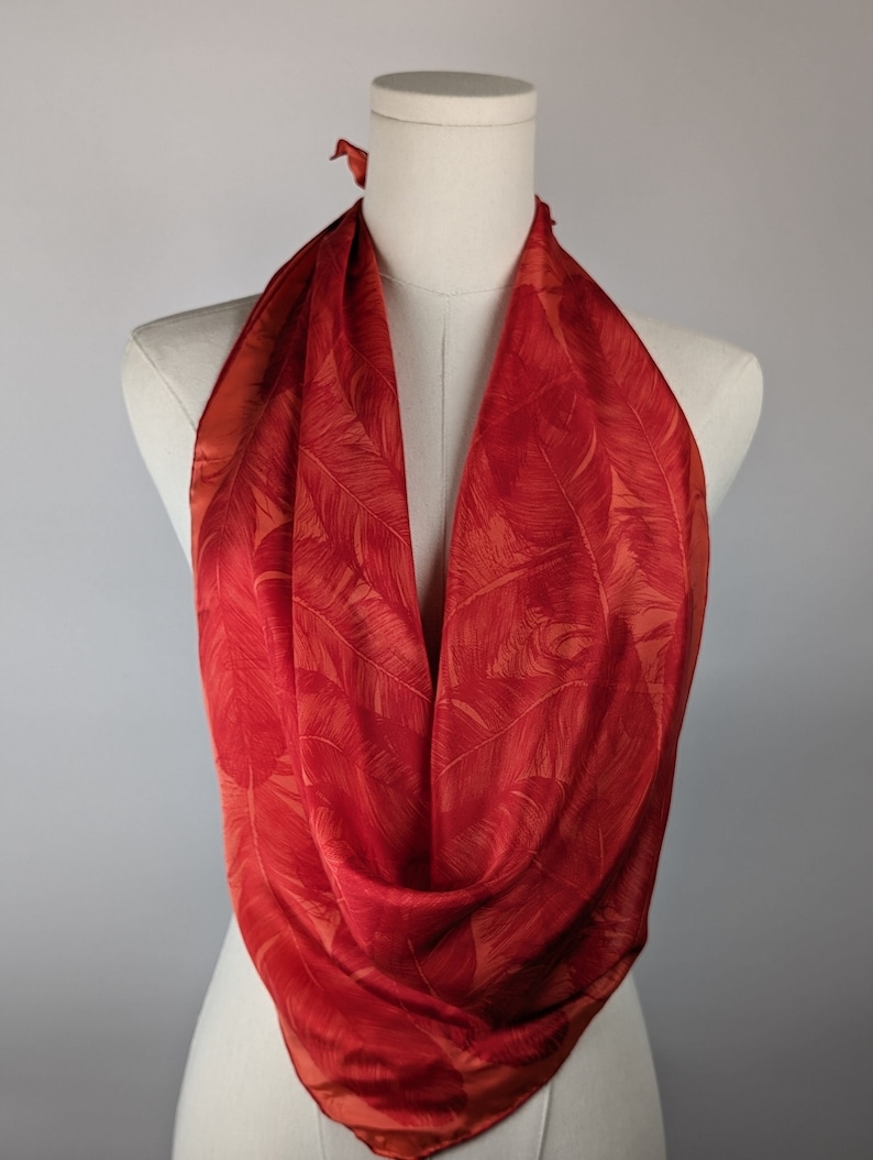 May include: A red silk scarf with a leaf pattern. The scarf is tied around a mannequin's neck.