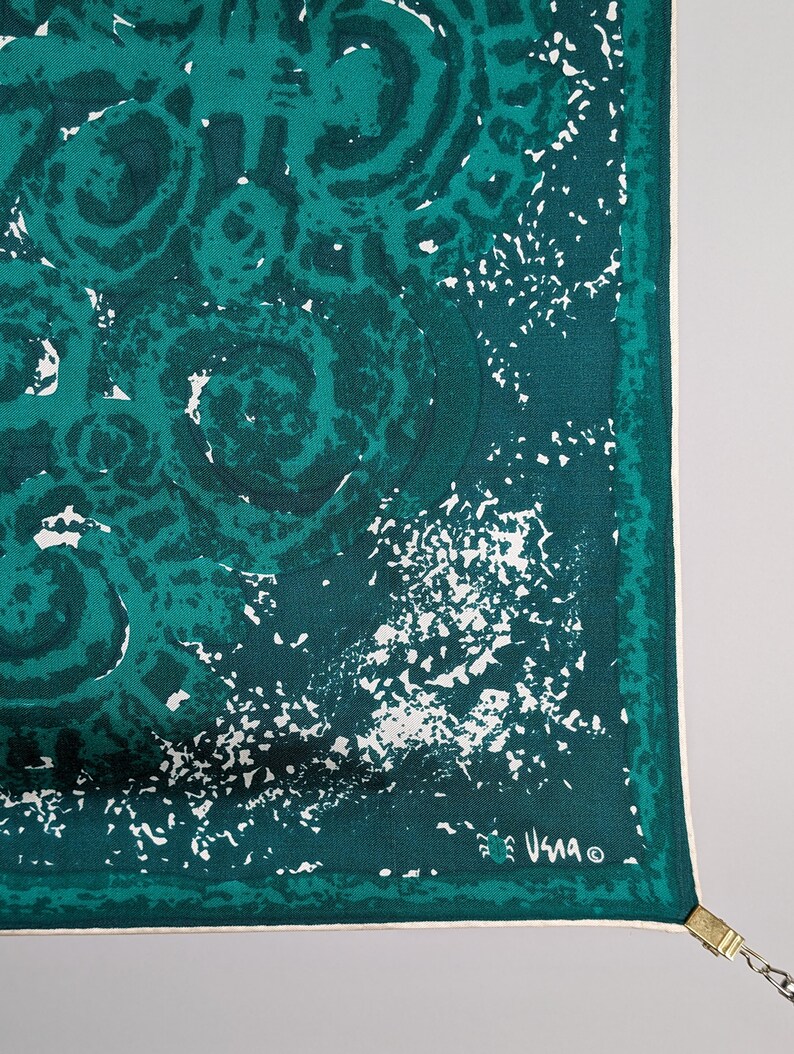 May include: A teal blue silk scarf with a white abstract pattern. The scarf has a white border and the word "Usma" is printed in the bottom right corner.