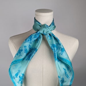 May include: A light blue silk scarf with a pattern of blue figures and a white background. The scarf is tied around a white mannequin's neck.