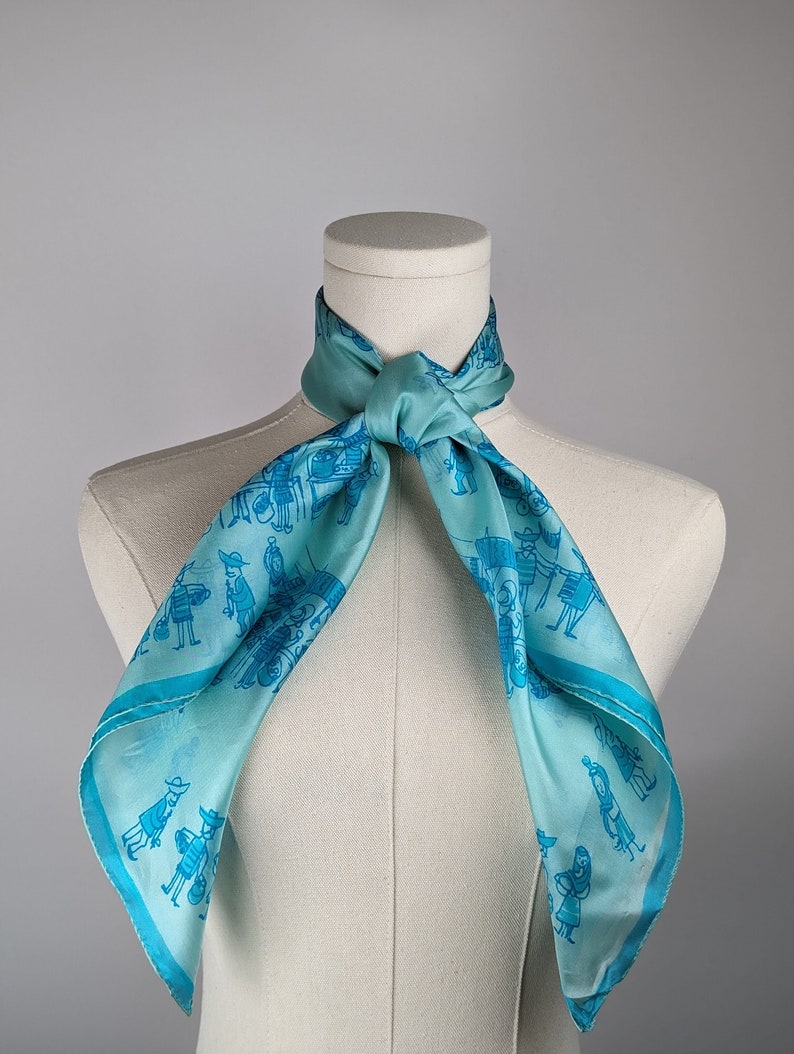 May include: A light blue silk scarf with a pattern of blue figures and a white background. The scarf is tied around a white mannequin's neck.