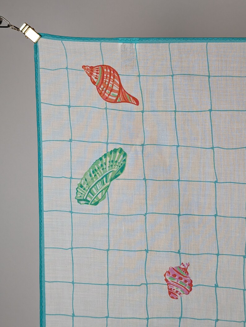 Op de afbeelding: Een wit katoenen zakdoek met een turquoise blauwe rand en een rasterpatroon. De zakdoek heeft drie kleurrijke schelpen in verschillende ontwerpen.