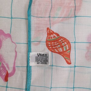 Op de afbeelding: Een wit katoenen zakdoek met een turquoise rand en een rasterpatroon. Het zakdoek heeft een herhalend patroon van roze en oranje schelpen. Het zakdoek heeft een label met de tekst "Una 100% Katoen 100% Coton Gemaakt in Japan Fait au Japon CA 04061 RN 25695".