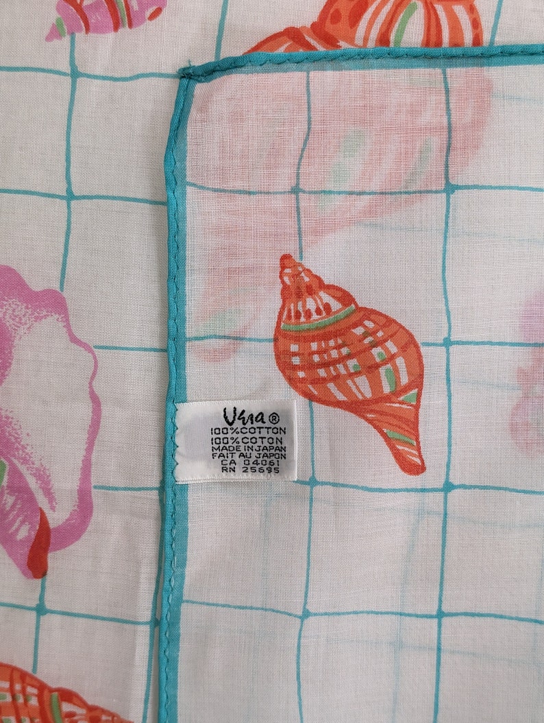 Op de afbeelding: Een wit katoenen zakdoek met een turquoise rand en een rasterpatroon. Het zakdoek heeft een herhalend patroon van roze en oranje schelpen. Het zakdoek heeft een label met de tekst "Una 100% Katoen 100% Coton Gemaakt in Japan Fait au Japon CA 04061 RN 25695".