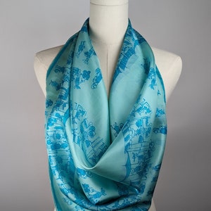 May include: A turquoise blue silk scarf with a repeating pattern of blue figures. The scarf is draped over a white mannequin.