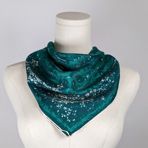 May include: A teal blue silk scarf with a white abstract pattern. The scarf is folded and displayed on a white mannequin.