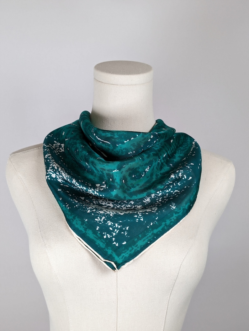 May include: A teal blue silk scarf with a white abstract pattern. The scarf is folded and displayed on a white mannequin.