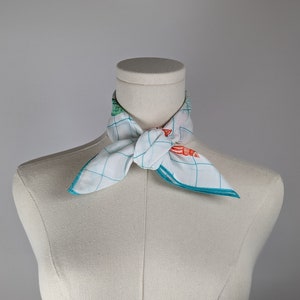 Op de afbeelding: Een witte sjaal met een blauw en groen geruit patroon en oranje en groene bloemenaccenten, gebonden om de nek van een wit mannequin.