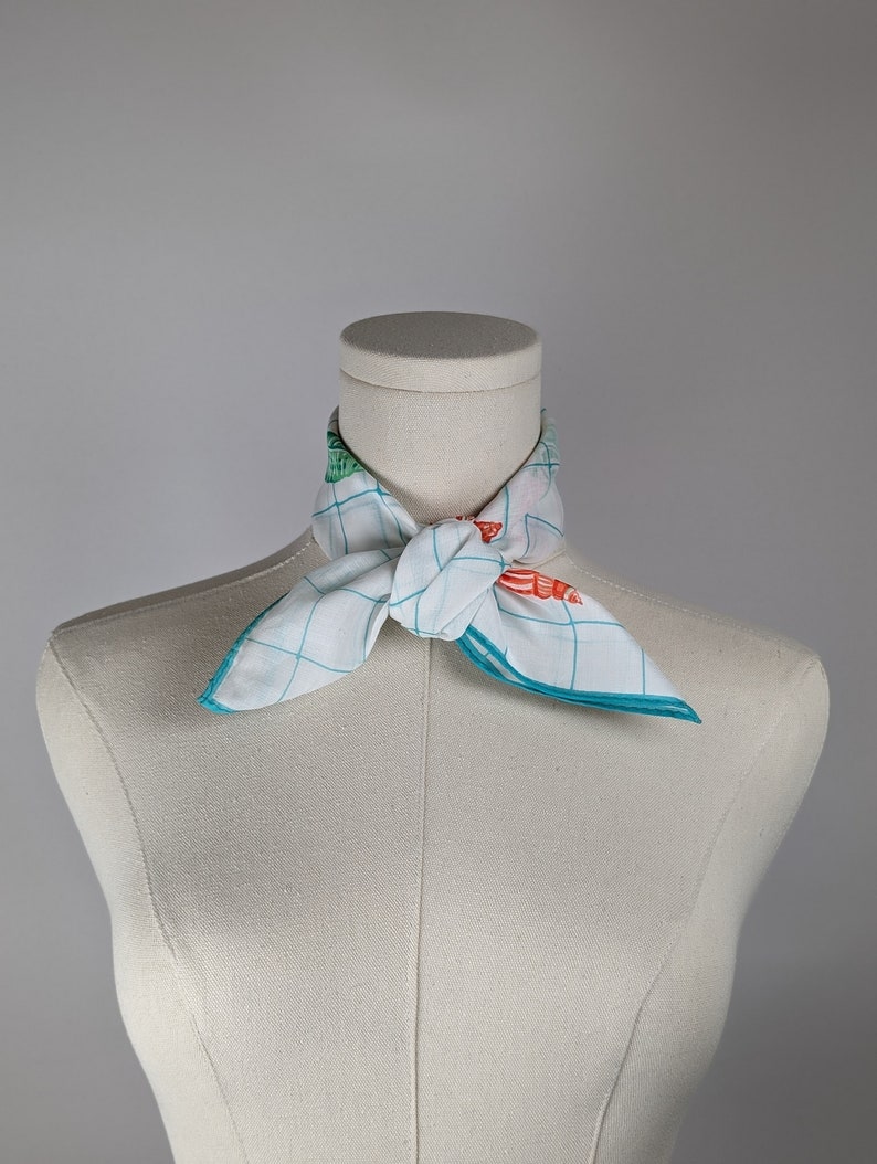 Op de afbeelding: Een witte sjaal met een blauw en groen geruit patroon en oranje en groene bloemenaccenten, gebonden om de nek van een wit mannequin.