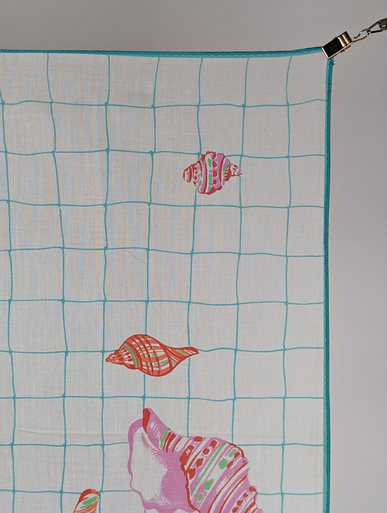 Op de afbeelding: Een wit katoenen zakdoek met een turquoise ruitpatroon en kleurrijke schelpontwerpen. Het zakdoek heeft een turquoise rand en is dubbelgevouwen.