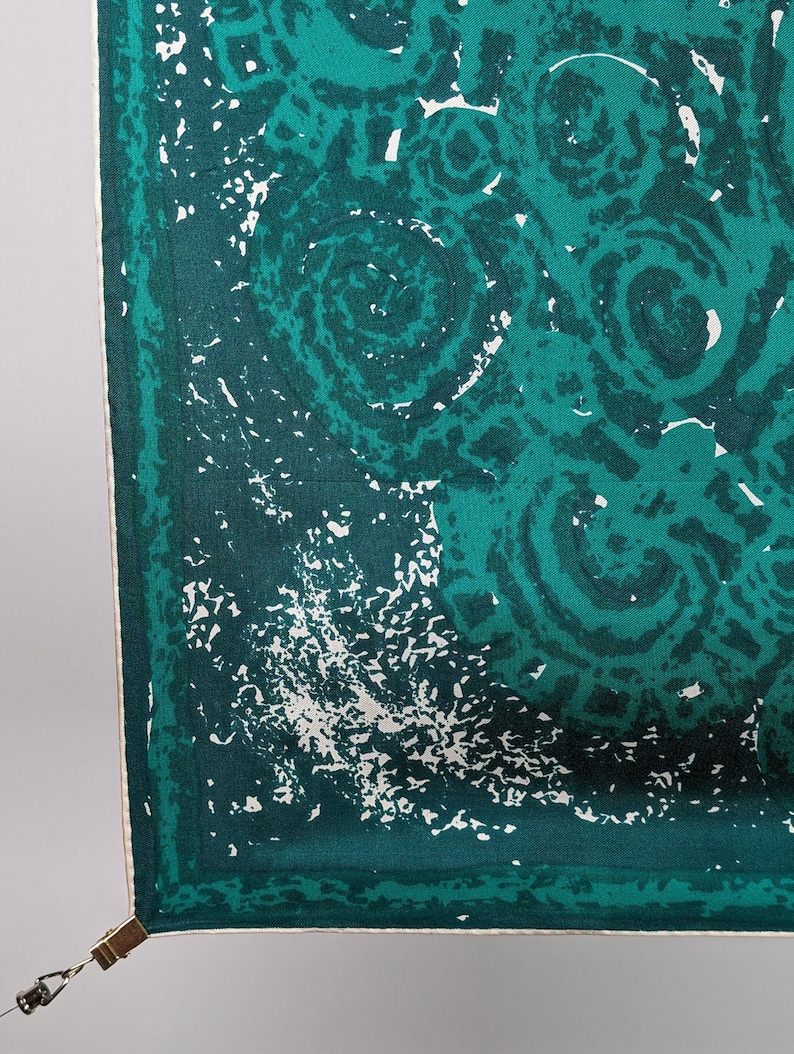 May include: A green and white patterned fabric with a white border. The fabric has a clip attached to the corner.