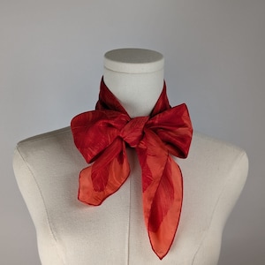 May include: A red silk scarf with a patterned design tied around a white mannequin's neck.